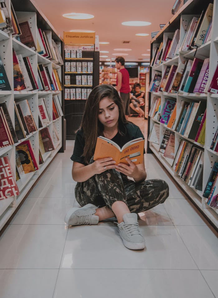 Young woman sitting on the floor and reading a book in a modern bookstore with shelves full of colorful books.